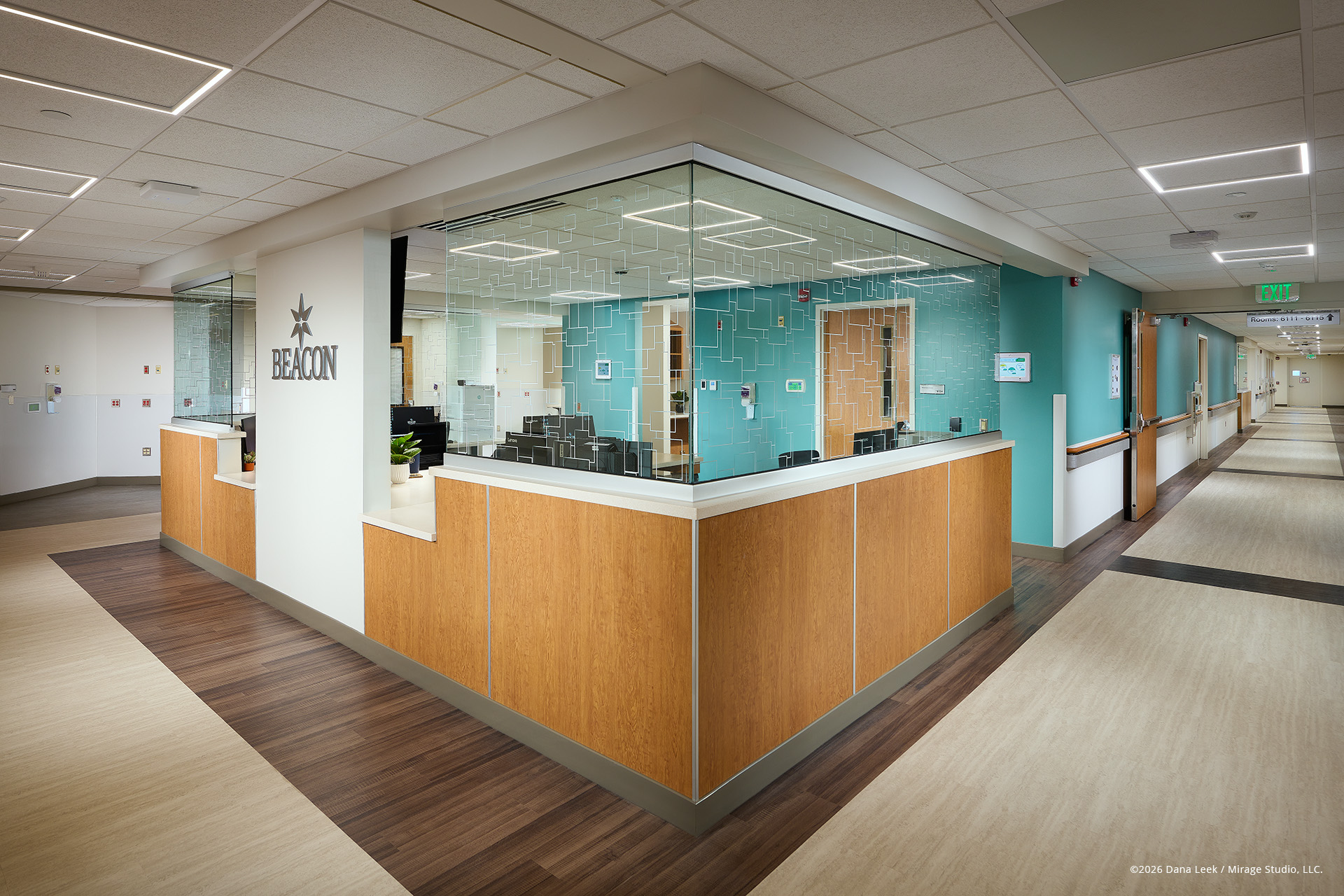 Beacon Health System nurse station at Elkhart General Hospital, with a glass‑wrapped work core and teal accent wall anchoring twin patient corridors. ​ ​