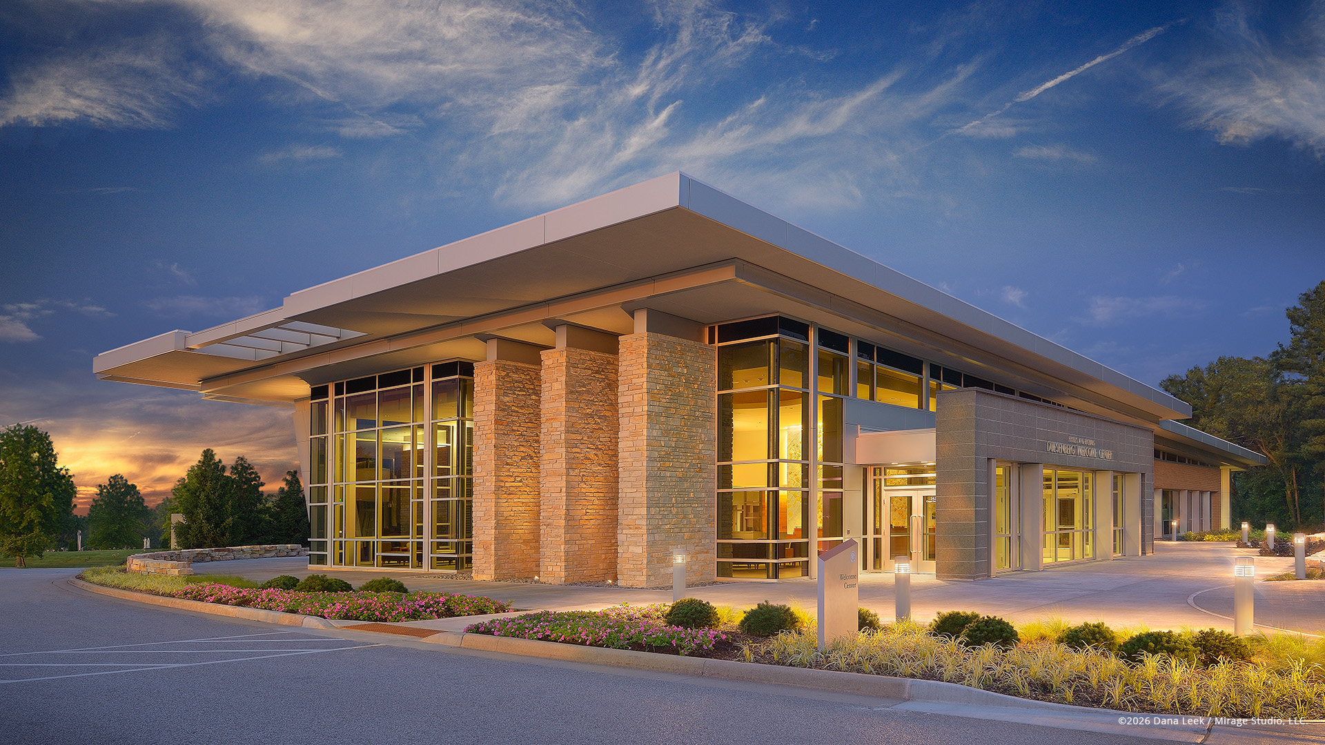 Valparaiso University’s Duesenberg Welcome Center glows at twilight, with warm interior light washing across its stone piers, glass curtain walls, and low, cantilevered roofline as sunset color lingers in the western sky.