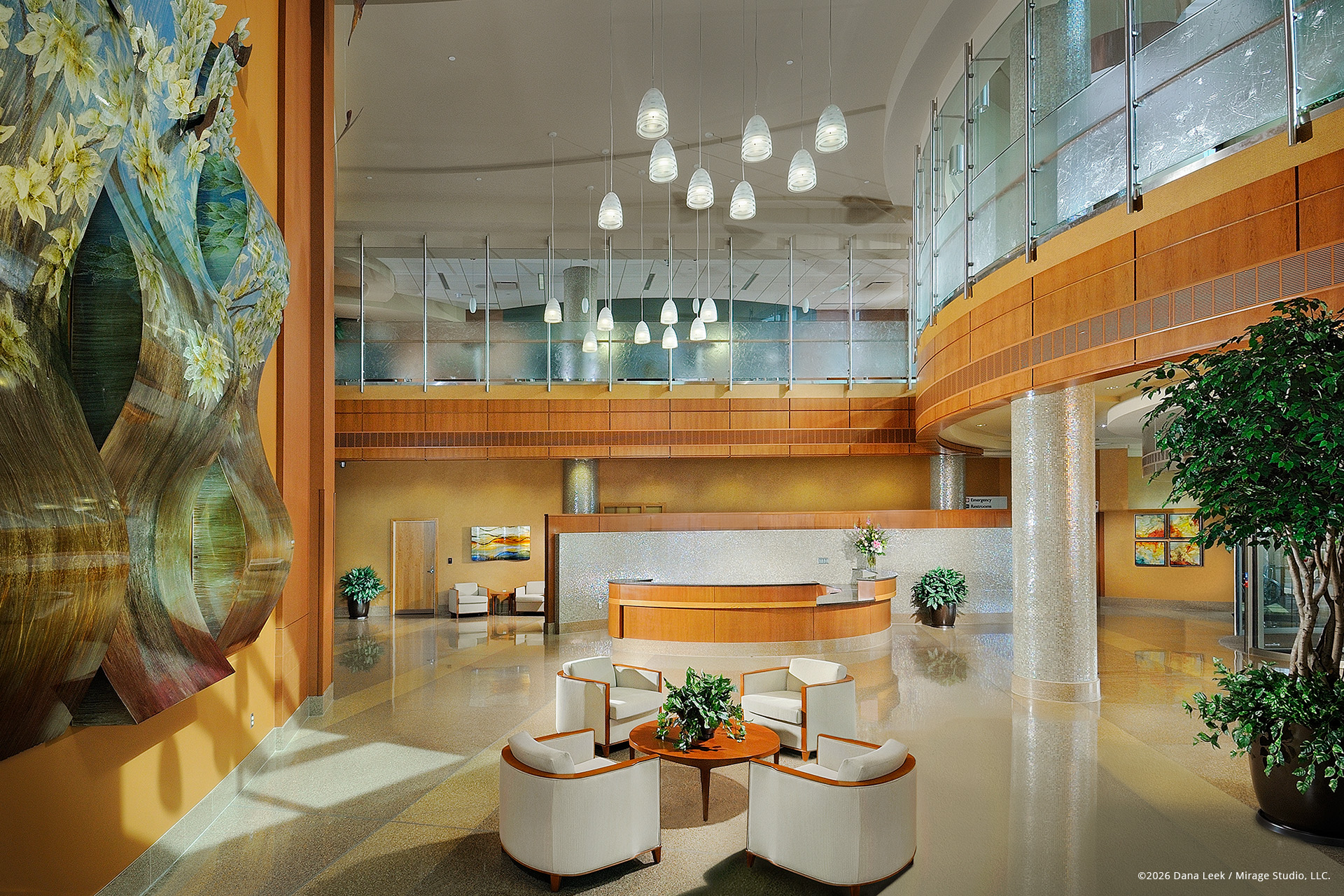 Two‑story hospital lobby with sculptural art wall, curved mezzanine, and concierge desk anchored by warm wood, terrazzo flooring, and cascading pendant lights.