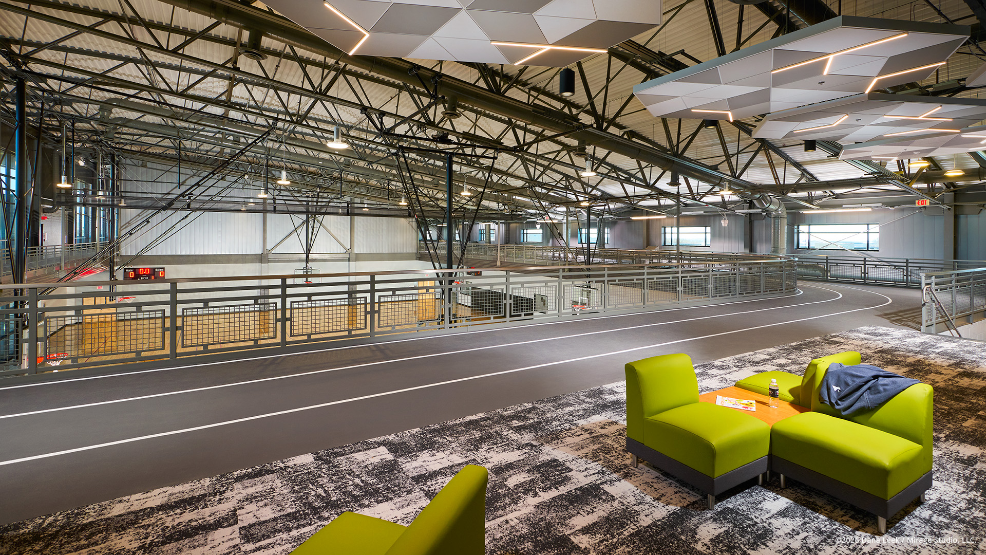Second‑floor lounge overlooking the community wellness center’s elevated indoor track and open multi‑court gym, framed by exposed steel structure and geometric acoustic clouds.