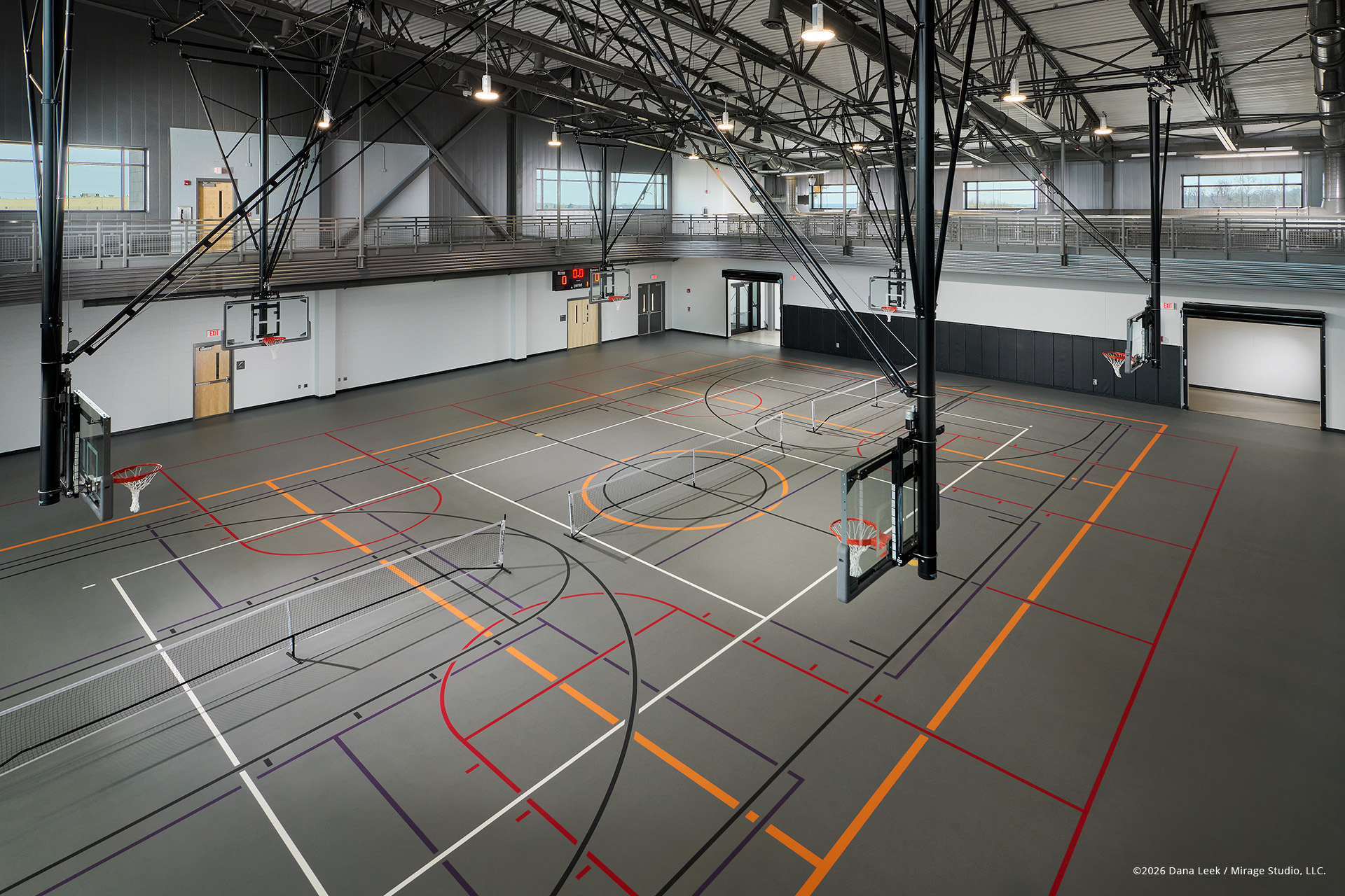 Multi‑court community gymnasium with retractable basketball goals, marked for basketball, volleyball, and pickleball beneath an elevated running track and exposed structure.