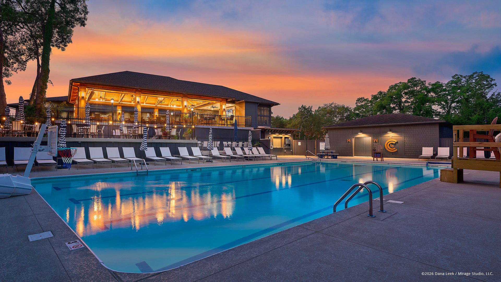 Long Beach Country Club’s pool deck glows in the soft light of sunrise, with the clubhouse lanterns reflecting in the calm water and rows of loungers lined up for a new summer day in Michigan City, Indiana.