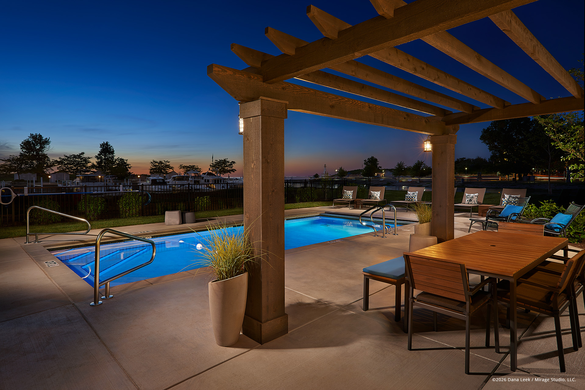 Twilight view of a lakeside residential pool and pergola terrace, with foreground architecture and seating lit by off‑camera strobes and landscape fixtures to balance the deep blue sky and glowing water beyond.