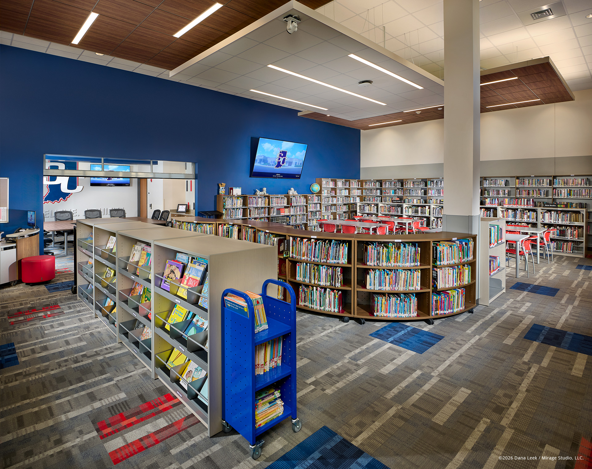 School library and media center with blue feature wall, low shelving for young readers, and round tables set among full‑height stacks and integrated technology.
