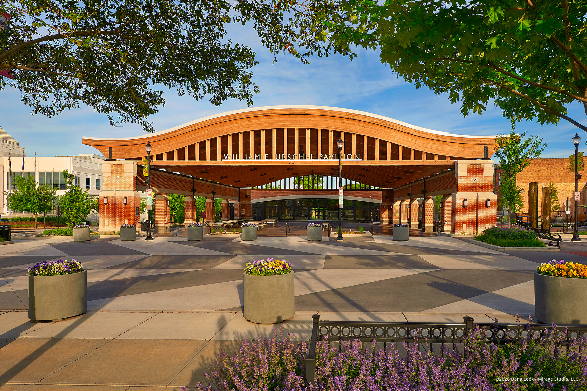 A welcoming summer view of the William E. Urschel Pavilion at Central Park Plaza in downtown Valparaiso, Indiana, with its signature arched timber roof, brick piers, and colorful planters framing the civic plaza.