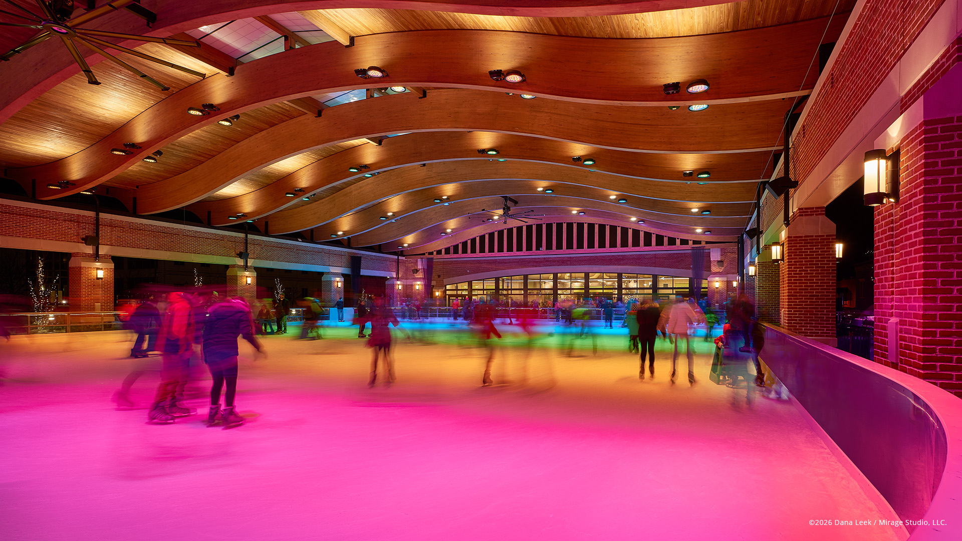 Color‑washed winter skating fills the William E. Urschel Pavilion in downtown Valparaiso, Indiana, with motion‑blurred skaters gliding under a sweeping timber roof and dynamic, color‑changing LED light.